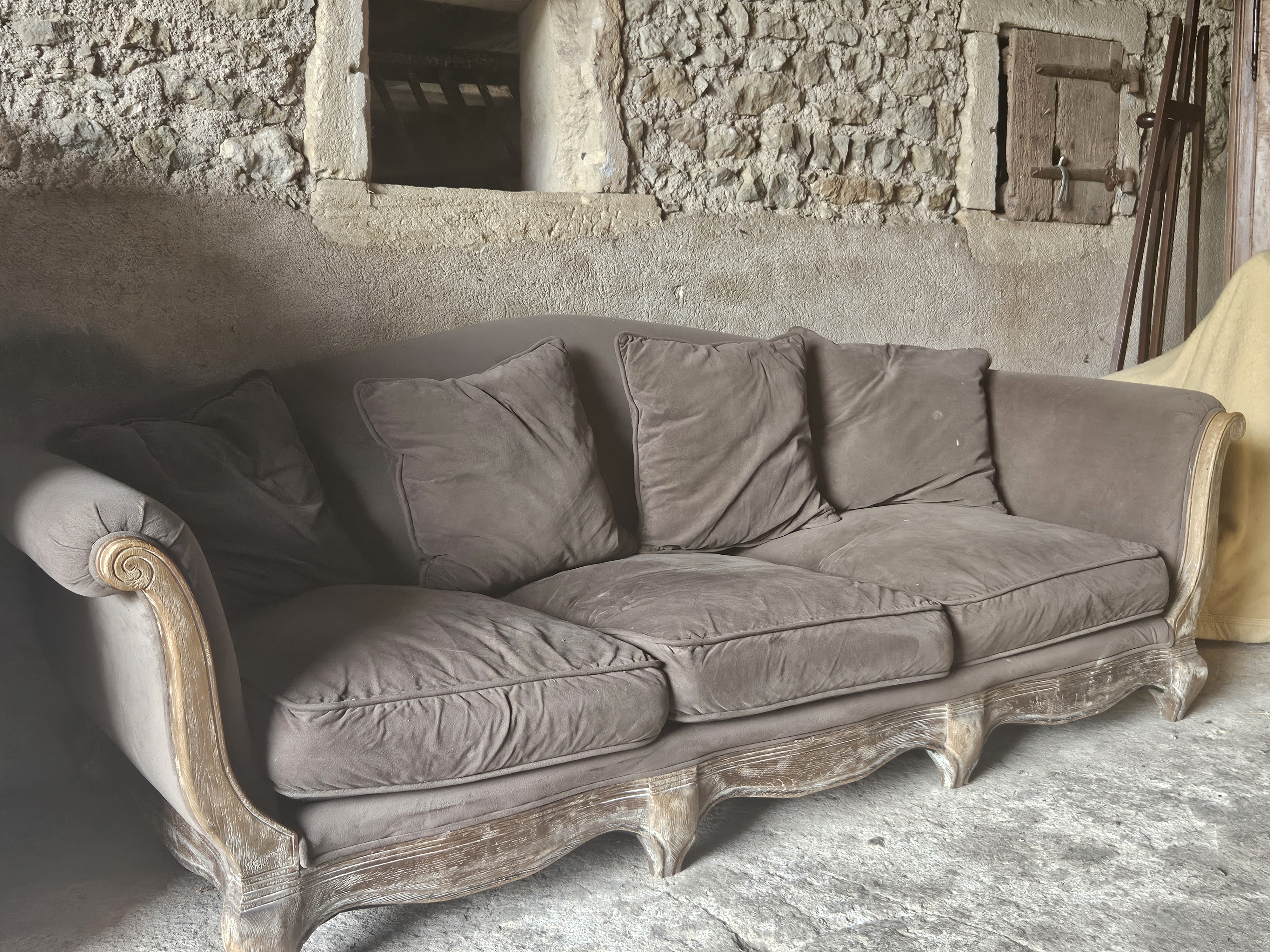 Canapé ancien gris à encadrement de bois clair sculpté dans un intérieur en pierre