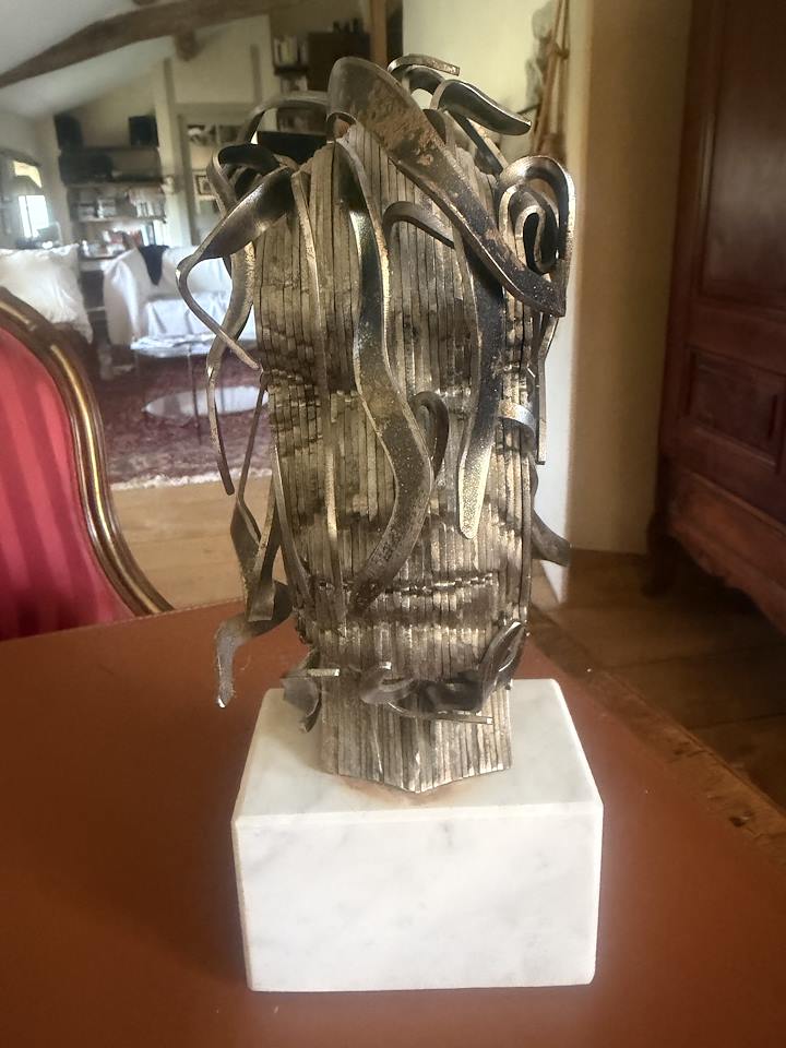 Sculpture abstraite argentée faite de lames et de courbes sur un socle blanc, vue de face